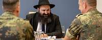 Ein Rabbiner mit schwarzem Hut unterhält sich an einem Tisch sitzend mit zwei Soldaten in Flecktarn