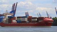 Ein Containerschiff mit gestapelten Containern an Deck. Im Hintergrund sind Kränen zu sehen.