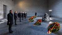 Boris Pistorius und weitere Personen stehen vor Blumenkränzen und einer Bronzeskulptur in einer Halle