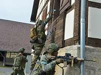 Ein Soldat auf einer Leiter wirft eine Handgranate in ein Fenster, zwei weitere sichern.