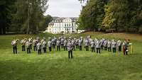 Das Heeresmusikkorps Kassel steht auf einer Wiese, im Hintergrund sieht man Schloß Wilhelmsthal 