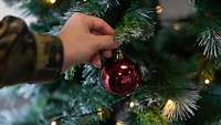 Eine rote Weihnachtskugel wird von einem Soldaten an einen Weihnachtsbaum gehängt.