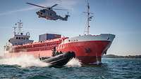Ein schwarzes Schlauchboot vor einem Containerschiff. Über ihnen ein Hubschrauber der Marine