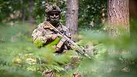 Ein Soldat steht mit seinem Gewehr zwischen mehreren Bäumen und Gräsern.