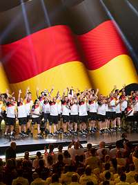 Mehrere Sportler stehen auf einer Bühne vor einer großen Deutschlandflagge und winken zum Publikum