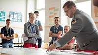 Ein Soldat zeigt auf einen Punkt auf einer vor ihm liegenden Karte hin. Um ihn herum stehen mehrere Zivilisten.