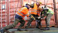 Drei Soldaten sichern einen Container an Deck eines Schiffes.