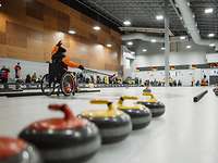 Eine Person in einem Rollstuhl zeigt mit einem Curlingstock auf einen Curling-Steine auf der Eisbahn