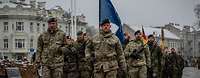 Ein Soldat marschiert an der Spitze einer Gruppe Soldaten. Hinter ihm weht die NATO Flagge. 