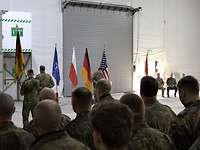 Mehrere Soldaten stehen in Formation. Im Hintergrund steht ein Soldat vor großen Fahnen an einem Rednerpult.