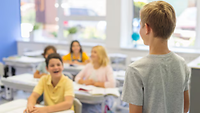 Ein Junge steht vor einer Klasse, mehrere Kinder sitzen an Tischen und schauen in seine Richtung.