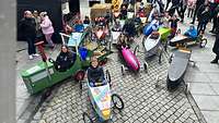 Blick von oben: Mehrere Kinder sitzen in bunt bemalten, verschieden geformten Seifenkisten.