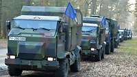 Several Bundeswehr vehicles are driving down a tree-lined road. Blue flags are attached to their right sides