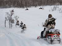 Soldaten im Schneetarnanzug fahren auf Motorschlitten auf einem verschneiten Feld in Richtung Wald.