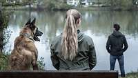 Eine Frau mit einem Hund links von ihr, blickt nachdenklich auf einen Mann der rechts neben ihr am Flussufer steht.