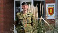 Eine Soldat mit schwarzem Barett steht vor einem Gebäude und verschränkt die Arme hinter dem Rücken.