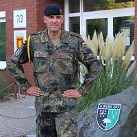 Ein Soldat mit goldener Kordel an der linken Schulter steht vor einem Gebäude.