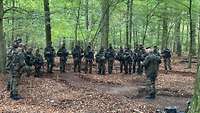 Soldaten stehen im Wald im Halbkreis um einen Soldaten, der zu ihnen spricht.