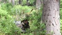 Eine Soldatin liegt gut getarnt zwischen Bäumen und Sträuchern im Wald.