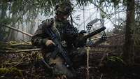 Ein Soldat mit einem Gewehr und einem Störapparat im Wald knieend.