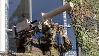 Zwei Soldaten stehen auf dem Balkon eines Gebäudes, einer trägt einen beigen Metallkasten mit Rohr.