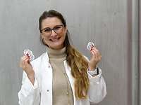 Eine Frau mit Brille trägt einen weißen Laborkittel und hält lächelnd fünf kleine Metallscheiben in die Kamera.