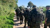 Eine Formation aus Soldatinnen und Soldaten mit zwei Reihen marschiert auf einem Waldweg.