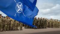 Soldaten verschiedener Nationen sind angetreten. Im Vordergrund wehen die Flaggen der EU und NATO.