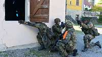 Soldaten knien vor einem Gebäude. Ein Soldat wirft eine Übungshandgranate in ein offenes Fenster.