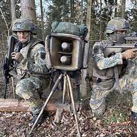 Zwei Soldaten knien im Wald zwischen einem Lautsprecher.