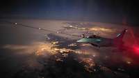 Ein Eurofighter bei Nacht fliegt über einer beleuchteten Landschaft und koppelt zum Betanken an.