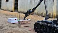 Ein Roboter mit Ketten und Greifarm setzt einen Störsender vor einem Paket in einer Gasse ab.