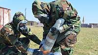 Zwei Soldaten mit ABC-Schutzanzügen und Masken packen ein Artilleriegeschoss in Folienbeutel.