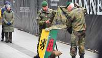 Zwei Soldaten entfernen von einem gold-grün-schwarz-roten Wappen eine Tarnfleckdecke.