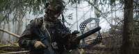 Ein Soldat mit einem Gewehr und einem Sensor im Wald knieend.