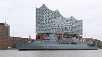 Ein graues Kriegsschiff liegt im Hafen vor einem Gebäude