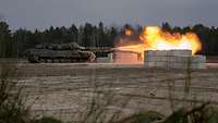 Ein Kampfpanzer schießt in Richtung aufgestapelter Betonsteine, ein Mündungsfeuer entsteht.