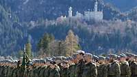 Soldatinnen und Soldaten stehen in einer Reihe, im Hintergrund sind Berge und ein Schloss zu sehen