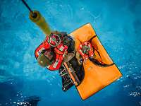 Eine Person mit Rettungsweste hängt an einem Seil. Unten im Wasser schwimmt eine Rettungsinsel.