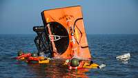 Personen in Schutzanzügen mit Schwimmweste und Helm schwimmen neben einer Rettungsinsel im Meer