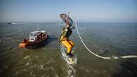 Eine Person im Schutzanzug mit Gurten springt an einem Seil in das Meer