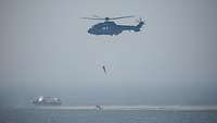 Ein Hubschrauber mit einer Person am Seil schwebt über einer Rettungsinsel und einem Boot im Meer