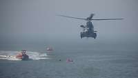 Ein Hubschrauber fliegt über das Meer, in dem ein Boot, Personen und eine Rettungsinsel zu sehen sind