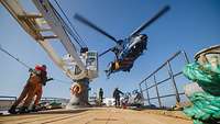 Ein Hubschrauber schwebt dicht über dem Deck eines Bootes, auf dem Personen stehen