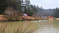 Mehrere Feuerwehren am Ufer eines Gewässers. Ein Unfallauto und Menschen im Wasser sind zu sehen.