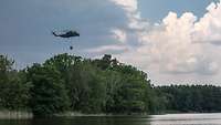Ein Hubschrauber fliegt über einem See, am Ufer stehen Bäume