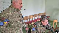 Ein Soldat aus Rumänien steht neben einem deutschen Soldaten, der sitzt. Beide schauen auf einen Laptop.