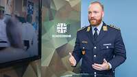 Ein Soldat steht an einem Tisch und spricht. Neben ihm steht ein Monitor.