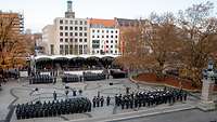 Angetretene Soldaten stehen auf einem Platz, im Hintergrund eine Tribüne mit Zuschauern und Gebäude
