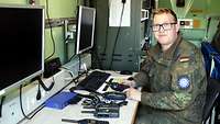 Ein Soldat sitzt an einem Tisch auf dem mehrere Funkgeräte liegen und schaut in die Kamera 